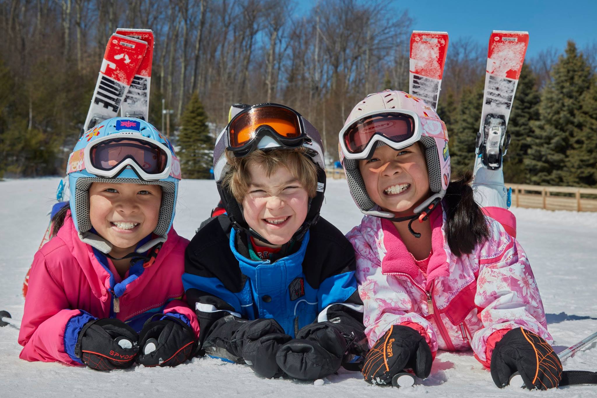 Three children laying on snow in skis, with goggles