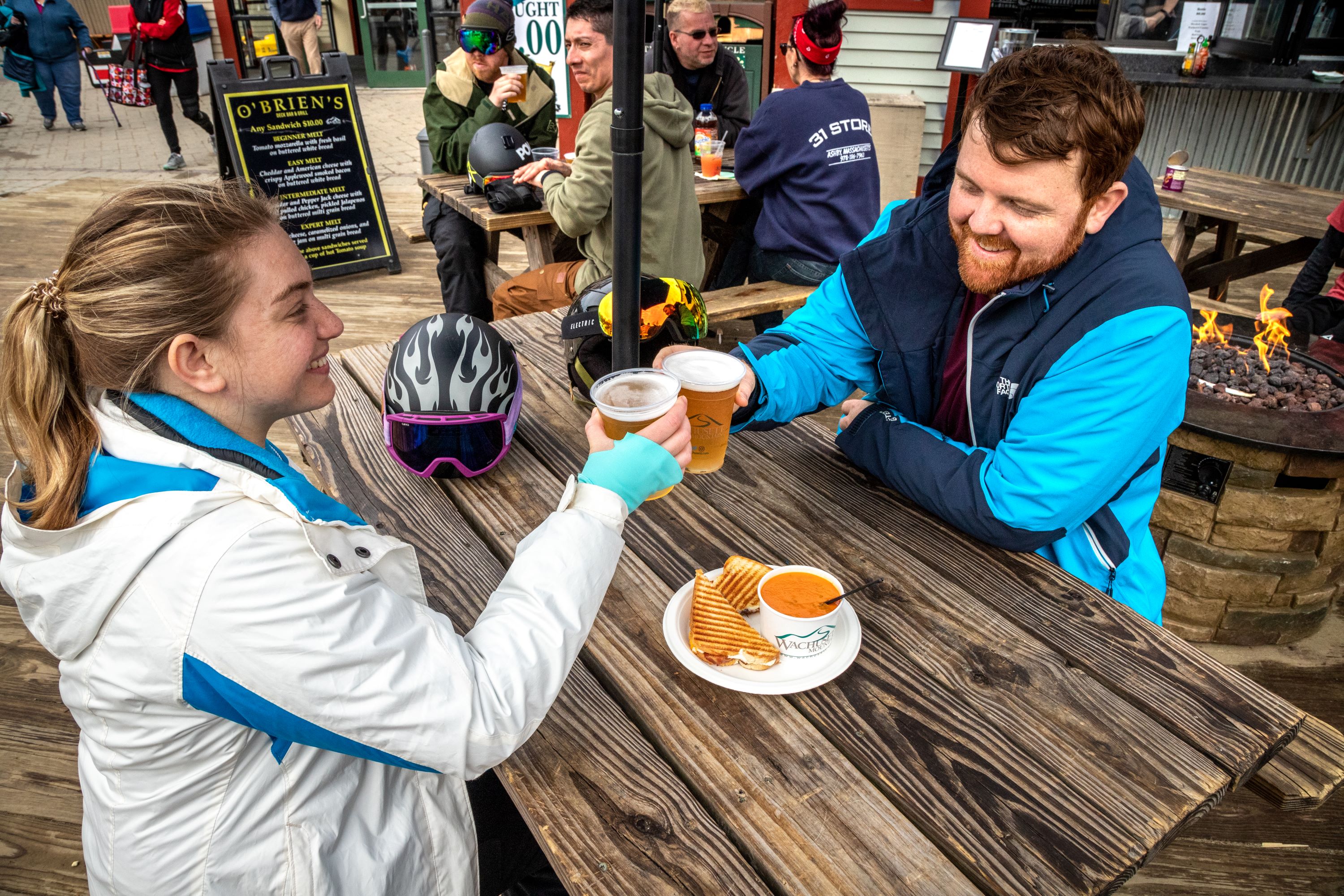 A woman in a white coat and a man in a blue coat clink glasses of beer sitting at a picnic table